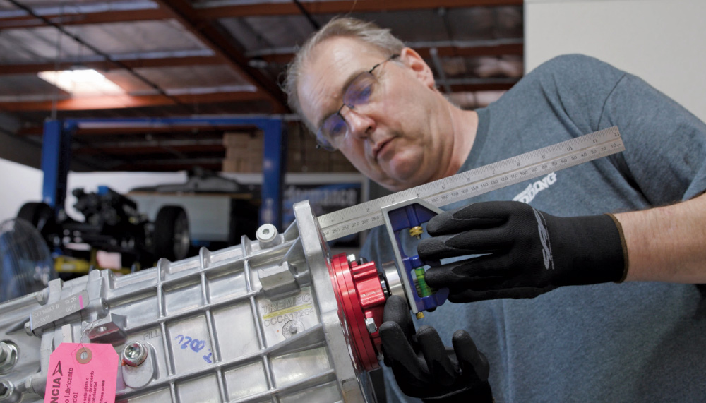 A mechanic wearing gloves and glasses uses a ruler to measure the position of the red hydraulic throw-out bearing on the front of a large, silver transmission.