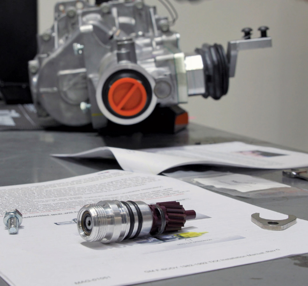 Close-up of a table showing a disassembled speedometer driven gear assembly and instructions, with a new silver transmission out of focus in the background.