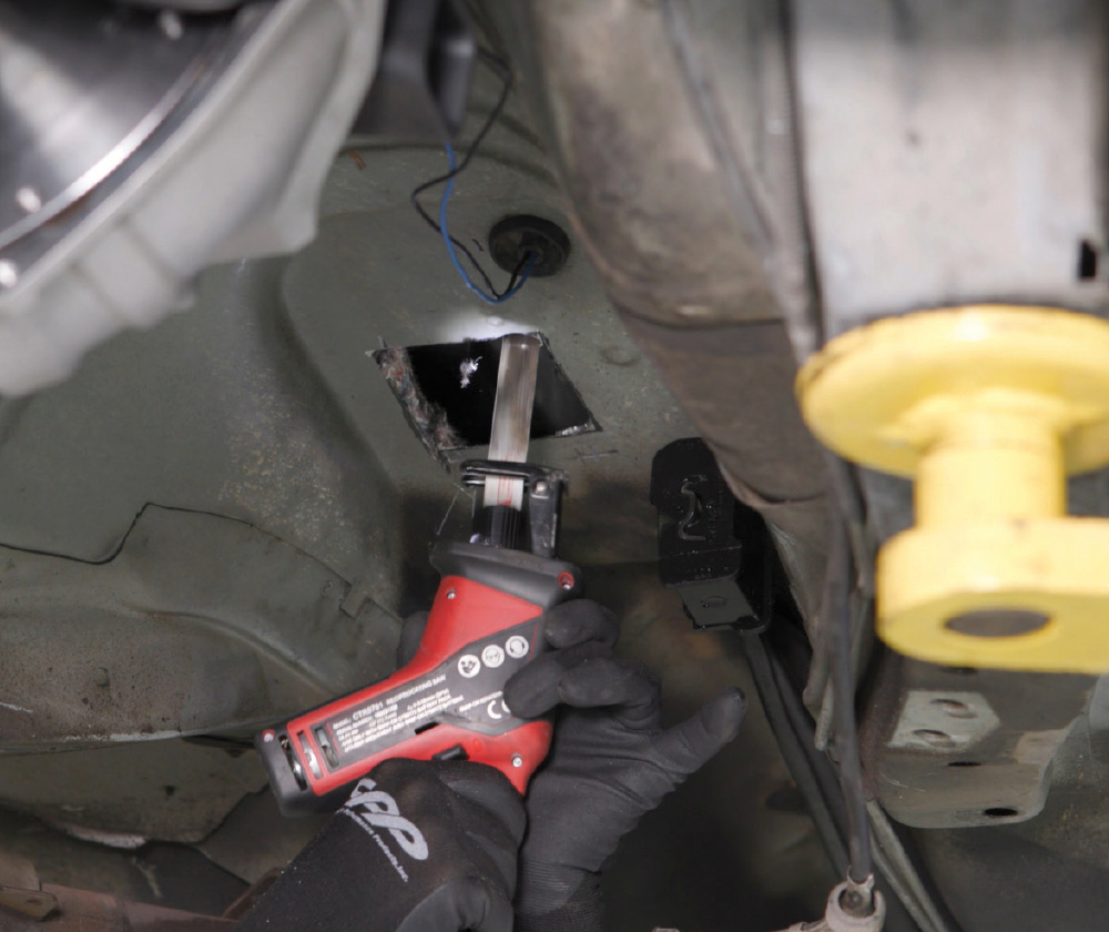 A mechanic uses a red reciprocating saw to cut a rectangular hole in the transmission tunnel floor of a car from underneath.