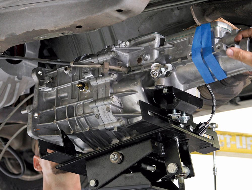 The new silver manual transmission on a black transmission jack is being guided into position underneath a raised vehicle.