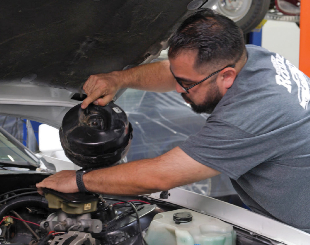 A mechanic with a beard and glasses is removing or installing the black brake booster from the engine bay of a white car.