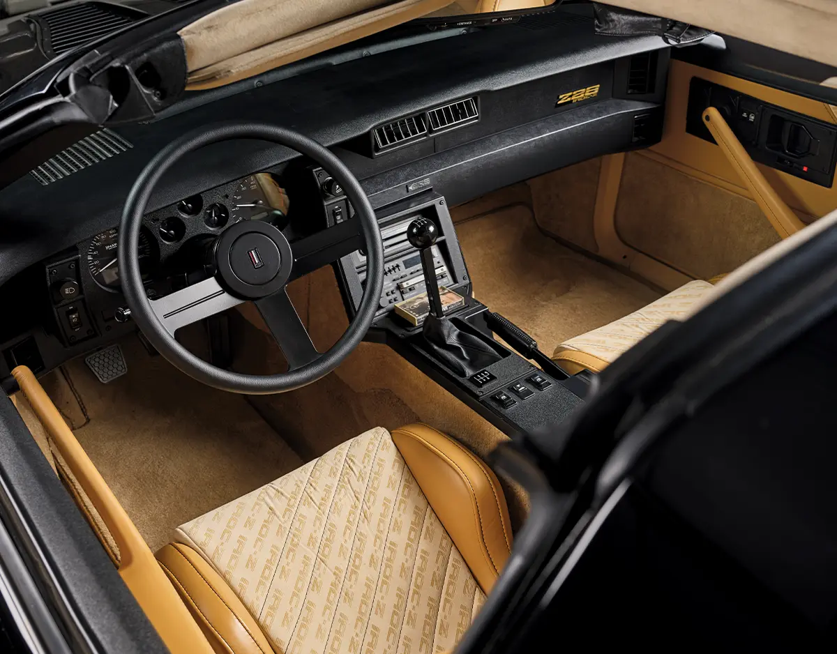 Interior view of a custom 1980s Chevrolet Camaro IROC-Z featuring a tan leather interior, integrated roll cage, and a manual shifter.