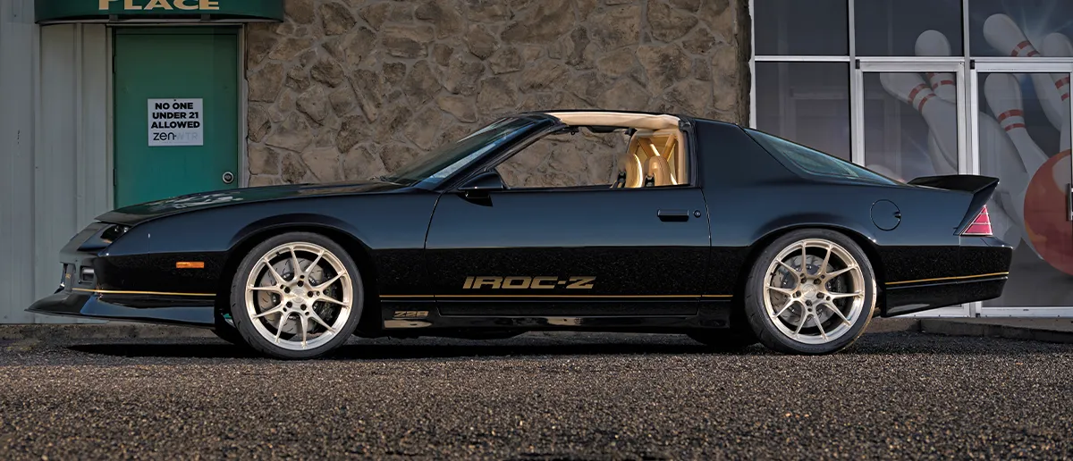A sleek black vintage third-generation Camaro IROC-Z featuring a tan interior roll cage, custom gold "IROC-Z" door decals, and high-performance brakes.