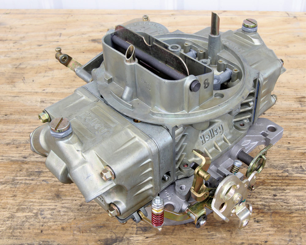 A top-down studio shot of a classic four-barrel Holley carburetor on a wooden workbench, showing the fuel bowls, throttle linkage, and gold-dichromate finish.
