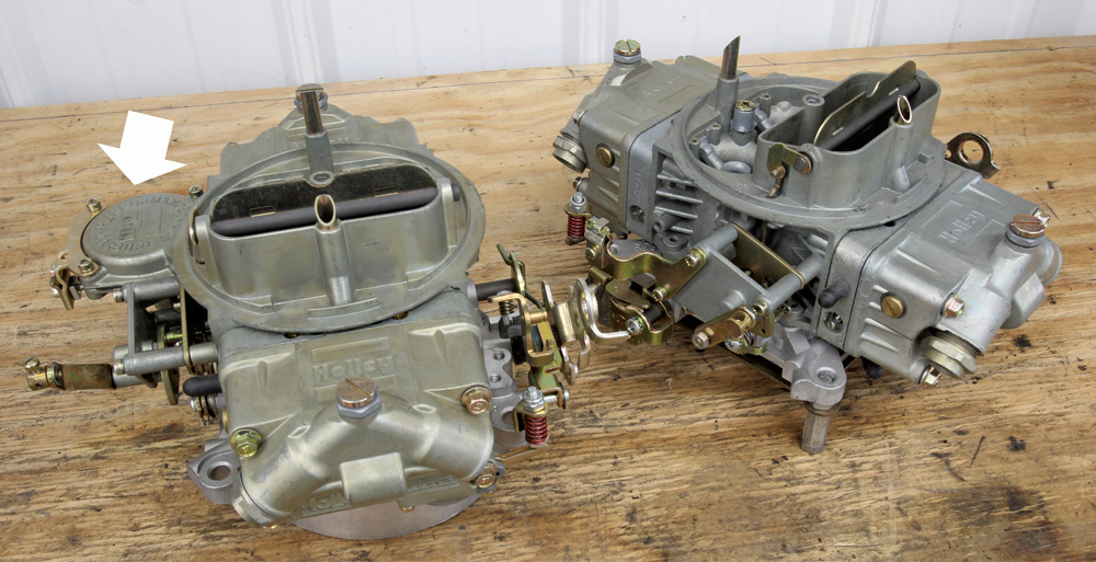 Two Holley carburetors placed side-by-side on a workbench, with a white arrow indicating the location of the vacuum secondary housing on the left unit.