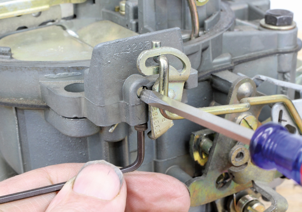 A close-up shot showing a technician using a flathead screwdriver and an Allen wrench to make fine adjustments to the carburetor's mechanical throttle linkage.