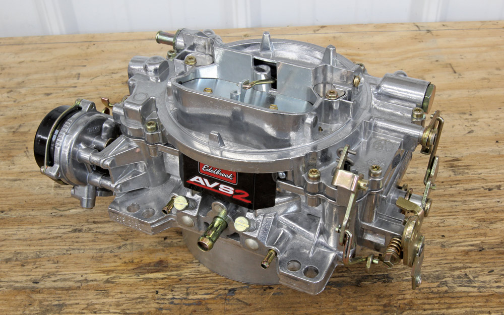 A detailed shot of an Edelbrock AVS2 carburetor on a workbench, showing its shiny aluminum finish, black branding label, and various vacuum port connections.