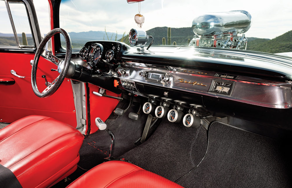 Interior of a 1957 Chevy featuring red leather bucket seats, a black dash with chrome accents, a Hurst floor shifter, and a large supercharger visible through the windshield.