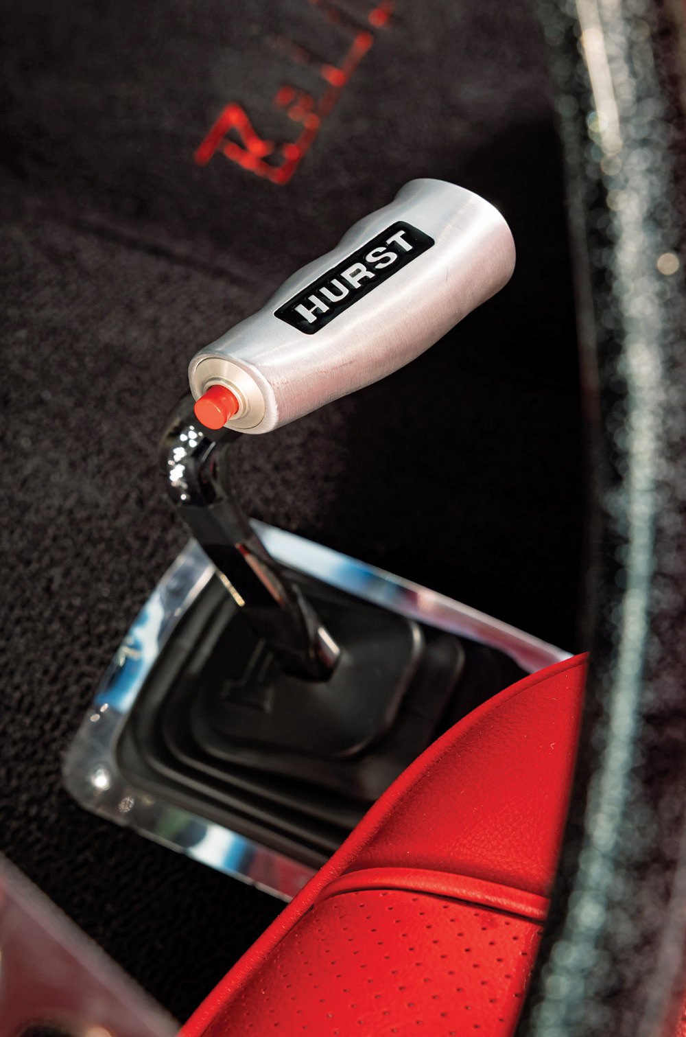 Close-up of a Hurst aluminum T-handle shifter with a red integrated button, mounted on a chrome lever with a black rubber boot and polished trim on black carpeting.