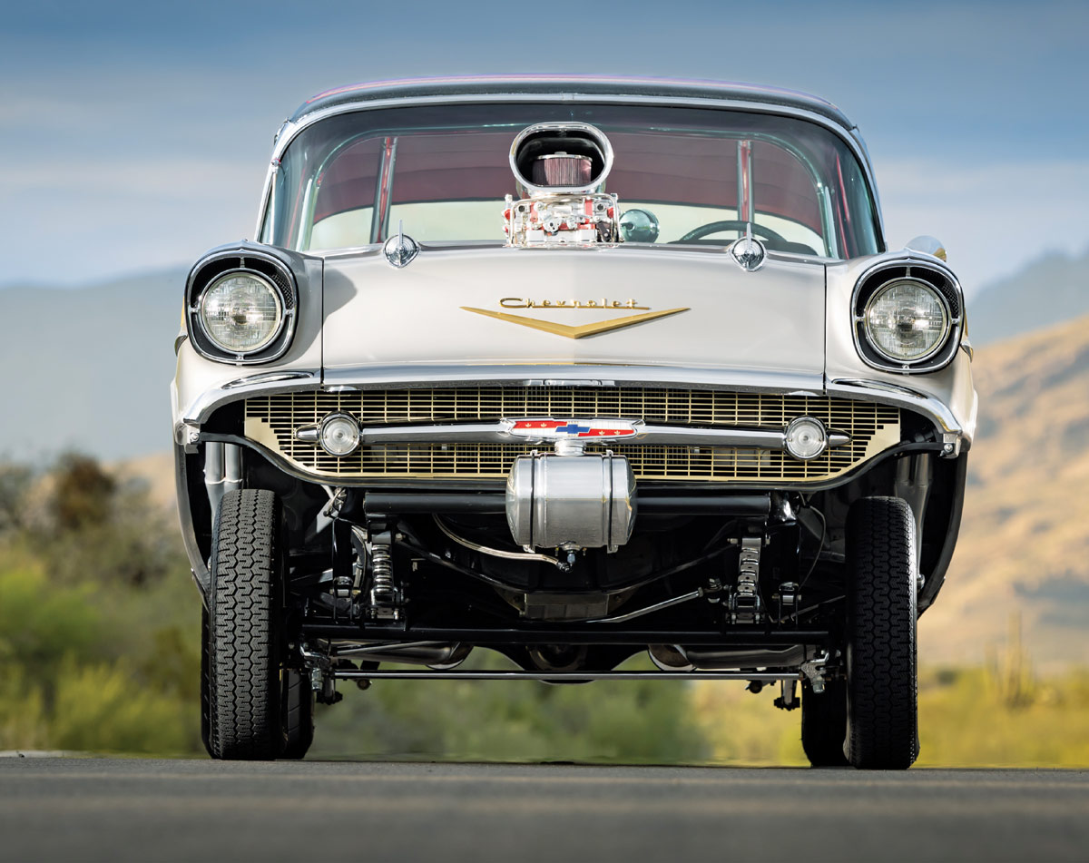 A low-angle head-on view of the 1957 Chevy gasser showing the moon tank, gold-colored grille, raised front suspension, and a massive blower intake dominating the hood line.