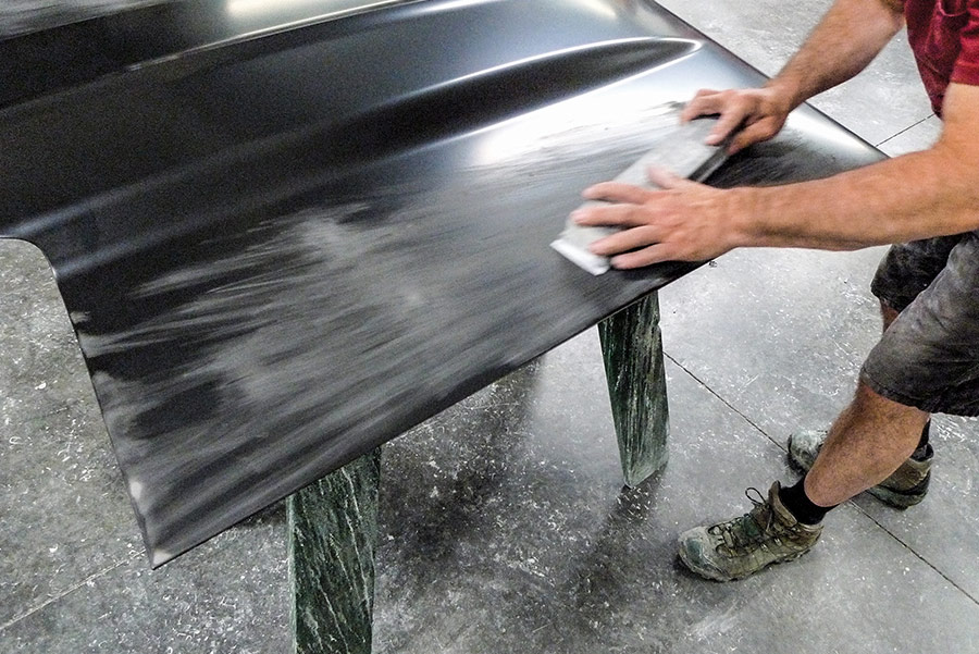 Technician hand sanding a new black primer-coated car hood on sawhorses as part of a classic car restoration project.
