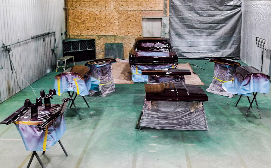 A classic maroon car and various body panels laid out on stands in a professional paint booth, showing the final stages of a full vehicle restoration.