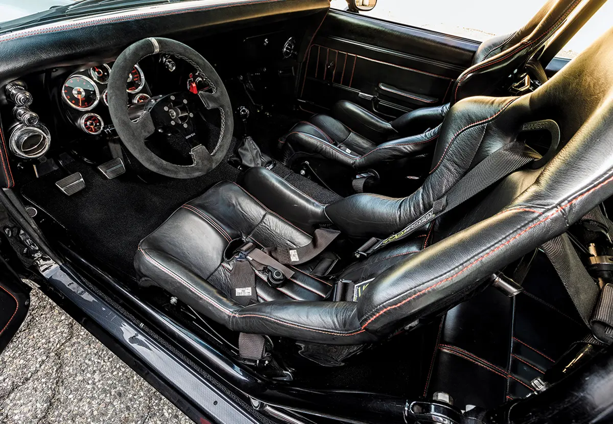 Interior of a black 1968 Camaro featuring two black leather bucket seats with red stitching and multi-point racing harnesses. A three-spoke suede-wrapped steering wheel faces a dashboard with multiple analog gauges.