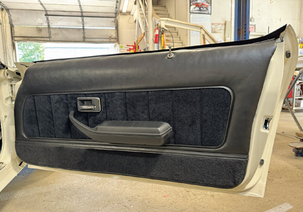 The finished black interior door panel is mounted on a white car door shell. It features a center cloth insert with vertical stitching, a black armrest, a chrome inner door handle, and a chrome door lock knob at the top.