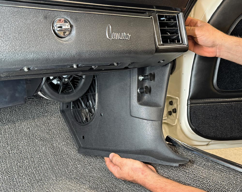 Hands positioning a black plastic kick panel in the passenger-side footwell of a Camaro. The dashboard above displays a "Camaro" emblem and air vent, while the floor is covered in grey looped carpet.