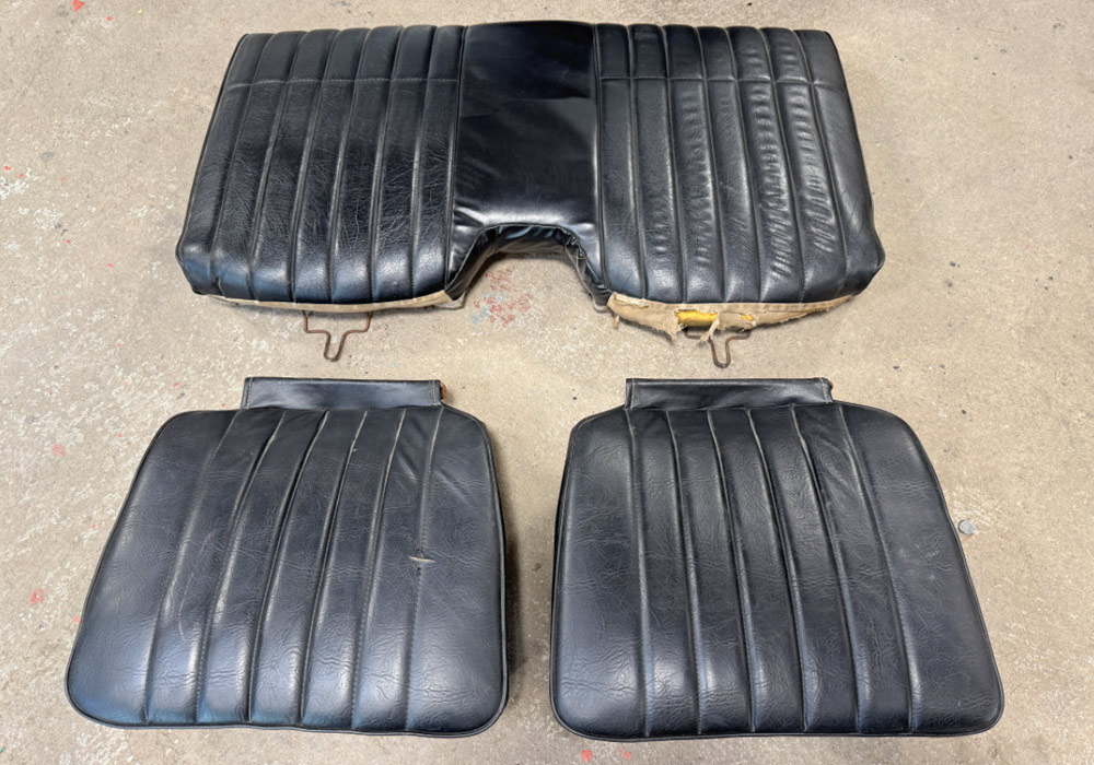 Three pieces of worn black vinyl rear seat upholstery with vertical pleats are laid out on a concrete floor. The set includes a large upper backrest section and two smaller lower seat cushions.