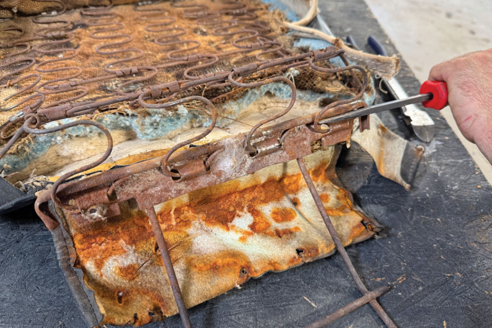 Close-up of a person using a flat-head screwdriver to separate the rusted metal inner support frame from the seat springs.
