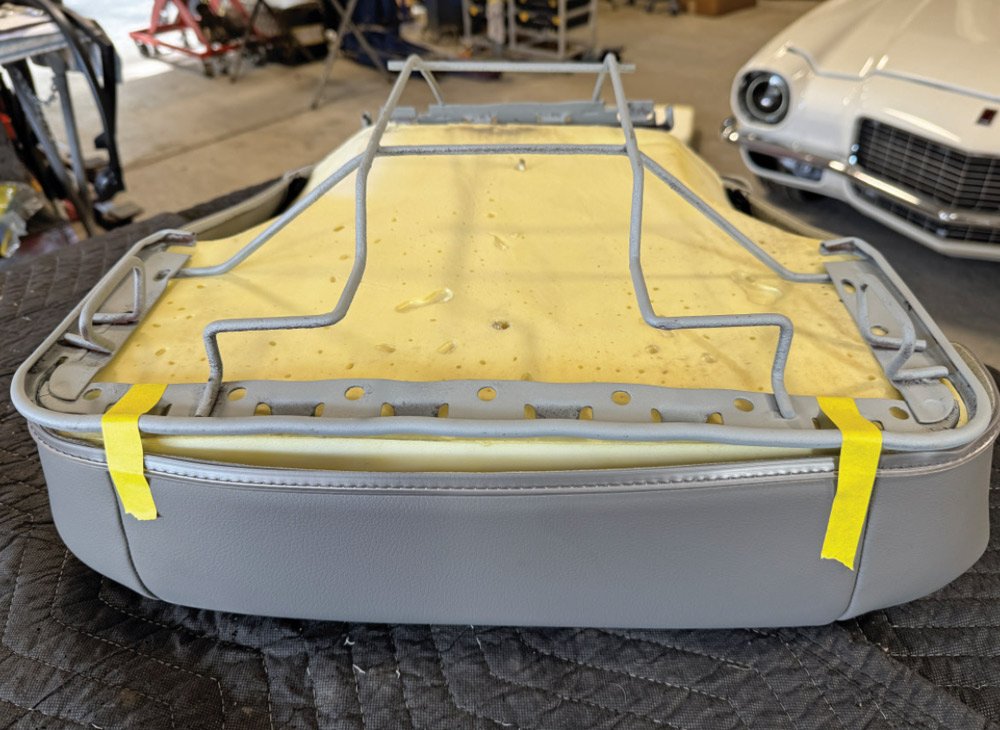 A grey car seat frame sits on yellow foam within an upside-down grey vinyl seat cover on a workbench. Two strips of yellow masking tape are placed on the front edge of the vinyl cover as alignment guides.