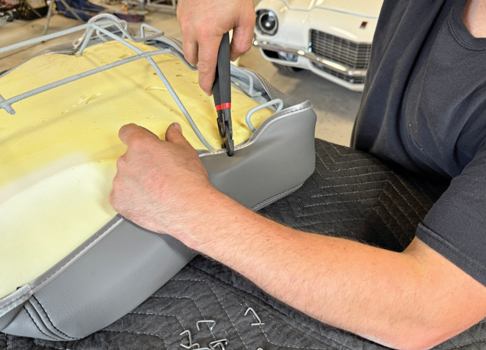 Hands use hog ring pliers to crimp a metal ring, attaching the edge of a grey vinyl seat cover to the metal wire frame on the underside of a seat cushion. Yellow foam is visible between the cover and the wire frame.
