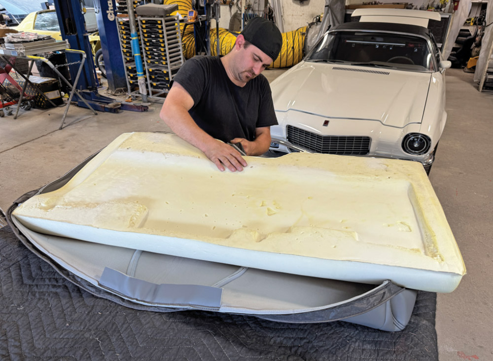 Pat O’Brien fits a large piece of white molded foam onto the back of a grey vinyl seat cover laid face down on a padded workbench. A white Camaro is visible in the background.