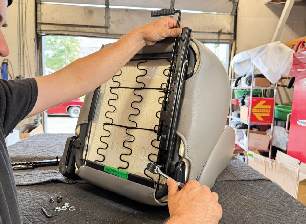 Hands use a socket wrench to bolt a black metal slider bracket to the bottom frame of a grey vinyl bucket seat. The seat is positioned upside down on a dark grey padded moving blanket.