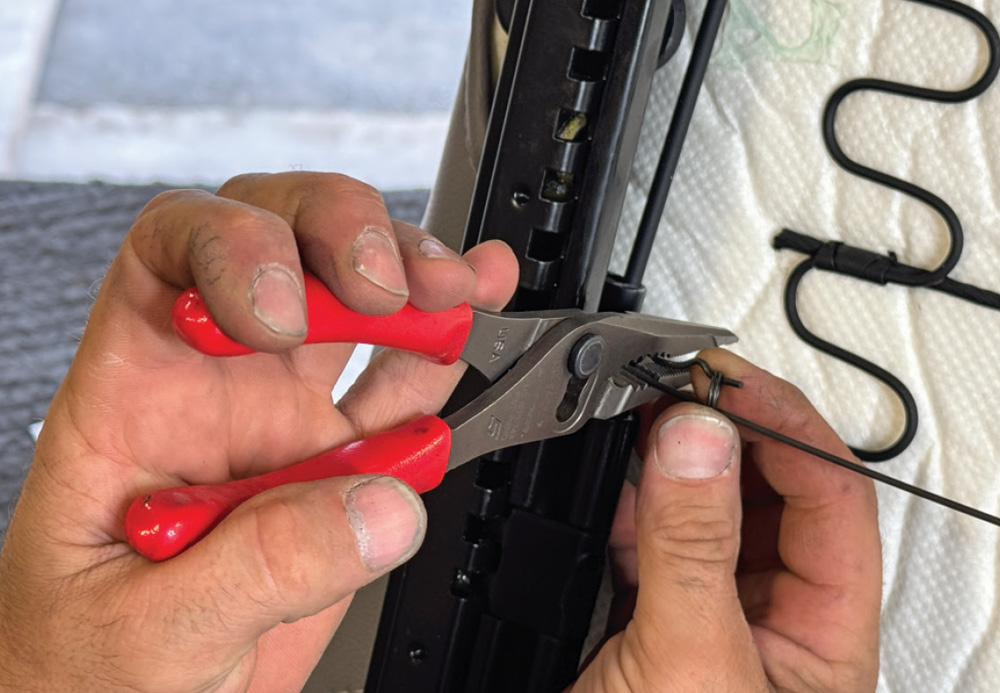 Close-up of hands using pliers to squeeze the ends of a thin wire rod together on a black metal seat slider track.