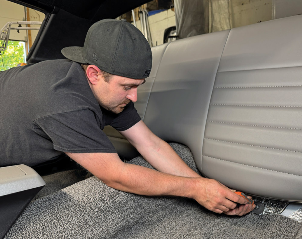 Pat O’Brien installs the grey vinyl rear seat backrest into the Camaro, using a hand tool to secure a bolt at the base near the grey carpet.