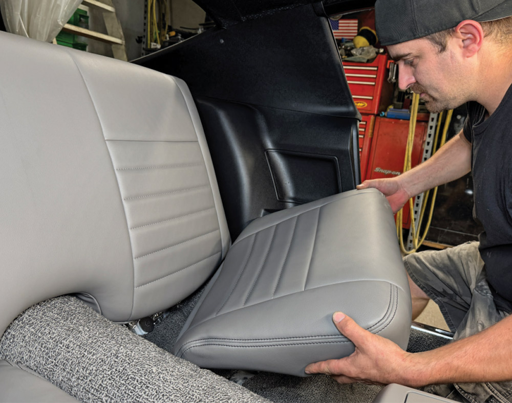 Pat O’Brien lowers a grey vinyl rear seat cushion into the passenger area of the Camaro, aligning it in front of the installed backrest.