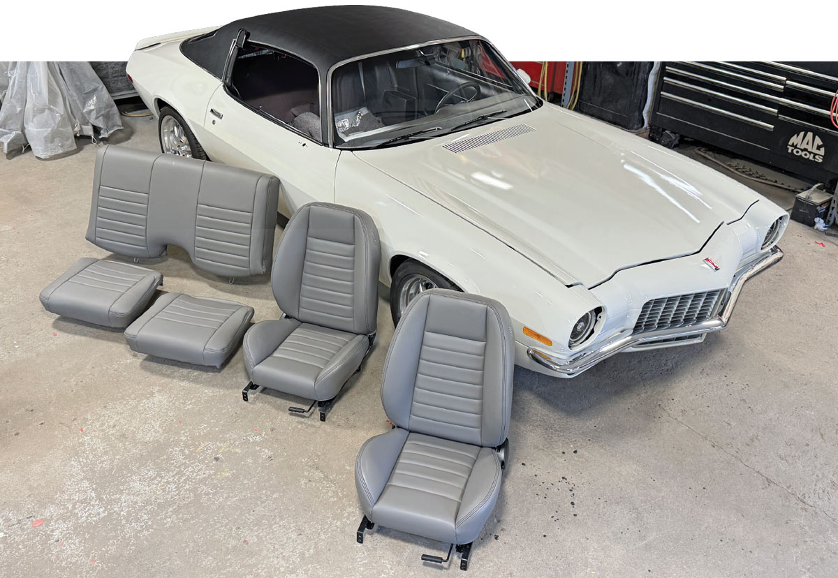 High-angle view of a white second-gen Camaro with a black vinyl roof parked in a garage. Two grey front bucket seats and two rear seat sections are laid out on the concrete floor in front of the vehicle.