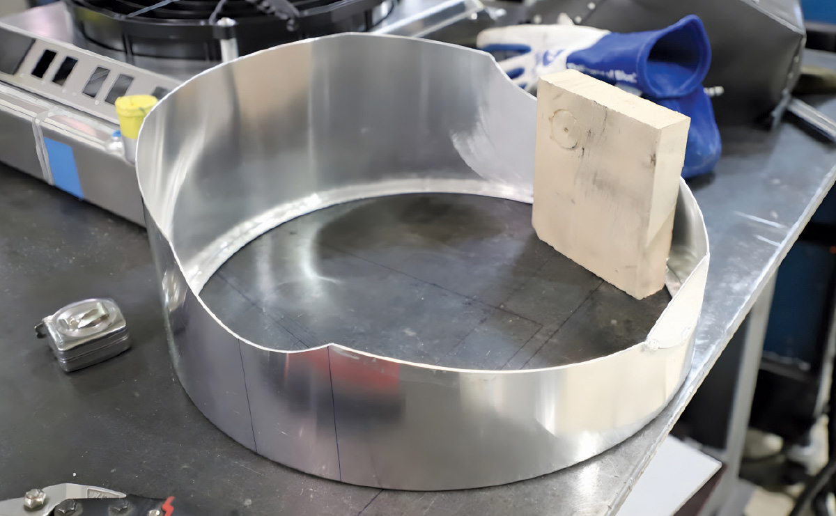 A curved aluminum sheet being fitted on a metal table next to a block of wood.