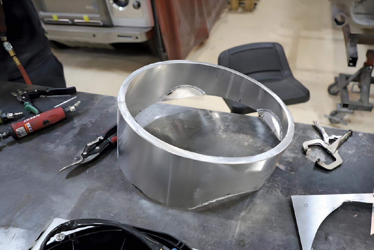 A welded aluminum circular shroud component resting on a workbench surrounded by tools.