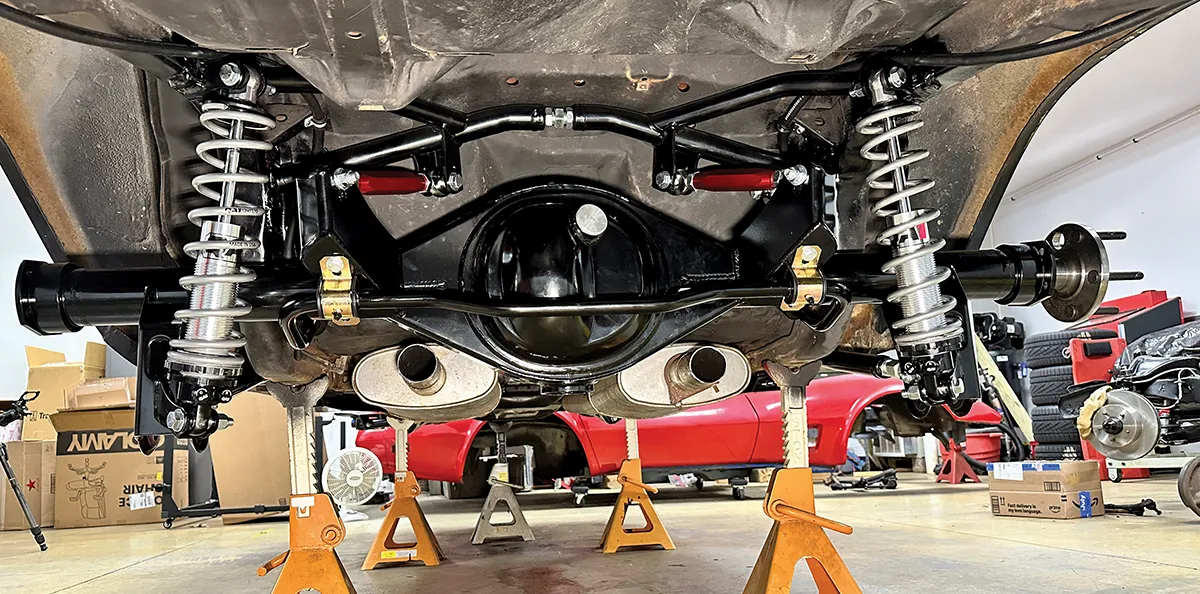 A high-performance rear suspension assembly on a classic Chevrolet Camaro, featuring silver coilover shocks, a black heavy-duty rear differential, and dual exhaust pipes, supported by orange jack stands in a professional garage