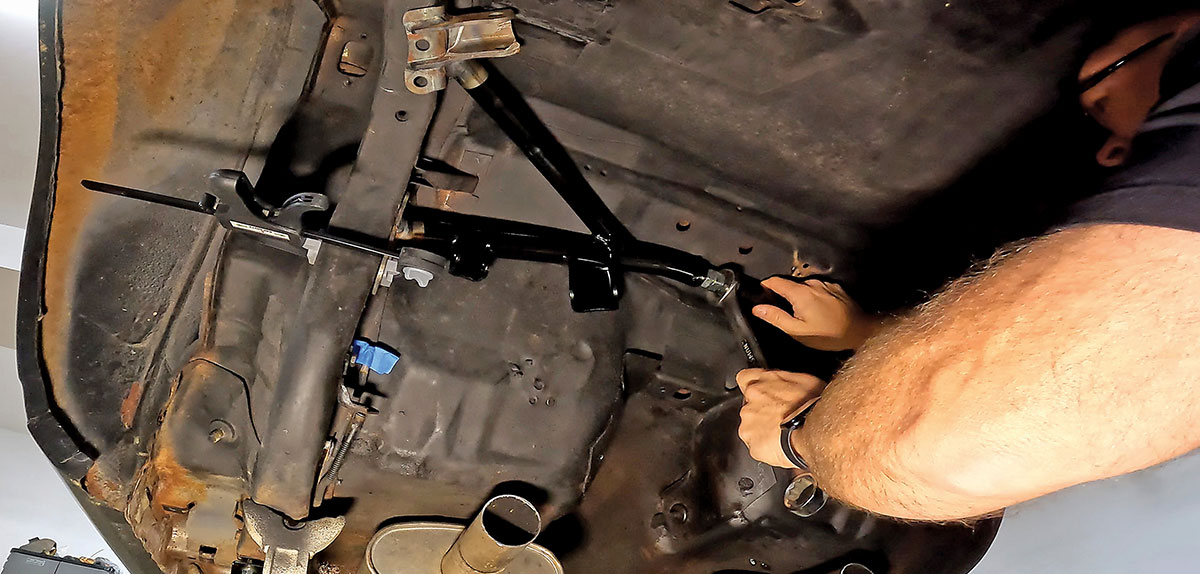 A mechanic installing a black powder-coated torque arm and chassis reinforcement bar on the undercarriage of a classic car project using a hand wrench