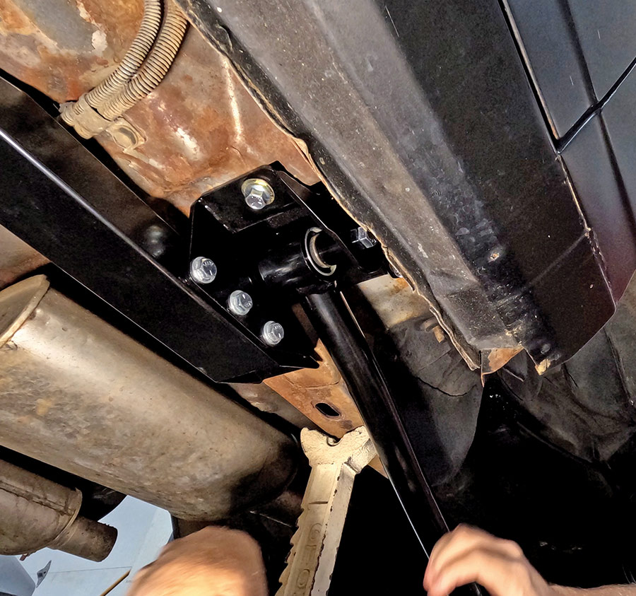 Installation of a heavy-duty black subframe connector on a classic car chassis to improve structural rigidity, featuring grade 8 mounting bolts and a silver jack stand for safety