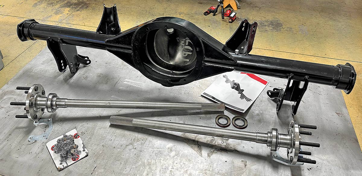 A dismantled high-performance rear axle assembly for a classic car project, featuring a black powder-coated differential housing, two long steel axle shafts with wheel studs, and new seals laid out on a workshop table