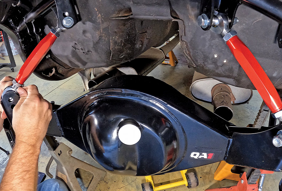 A mechanic installing a heavy-duty QA1 Ford 9-inch rear axle housing into a classic car, connecting it to adjustable red powder-coated torque arms and a custom chassis crossmember