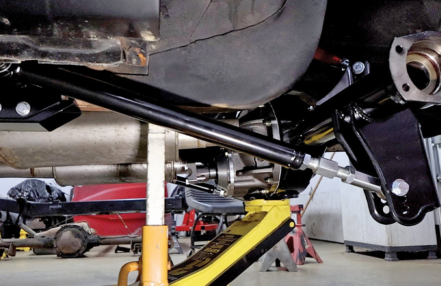 A side view of a pro-touring suspension installation on a classic car, featuring a black torque arm connected to a Ford 9-inch rear axle housing and supported by a yellow floor jack