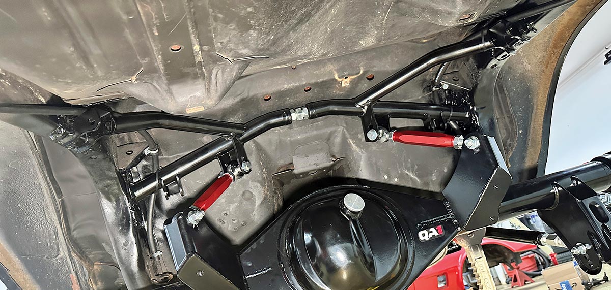 A wide-angle view of the completed QA1 rear suspension conversion on a classic car, showing the Ford 9-inch axle housing, red adjustable torque arms, and black chassis reinforcement crossmember