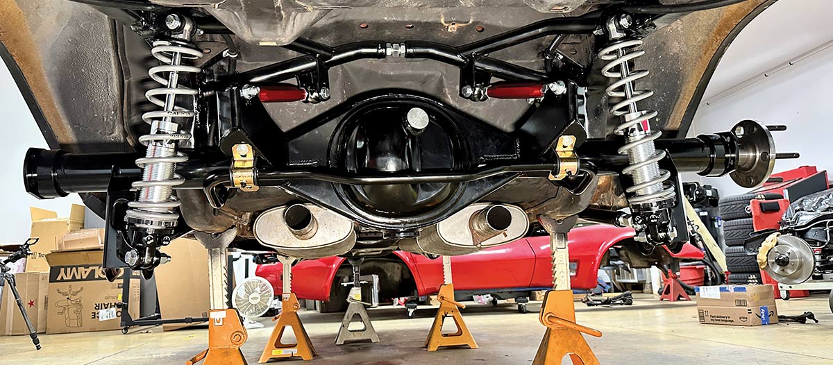 A wide-angle undercarriage view of a completed QA1 rear suspension conversion on a classic car, featuring a black Ford 9-inch axle, silver adjustable coilover shocks, red torque arms, and a heavy-duty sway bar