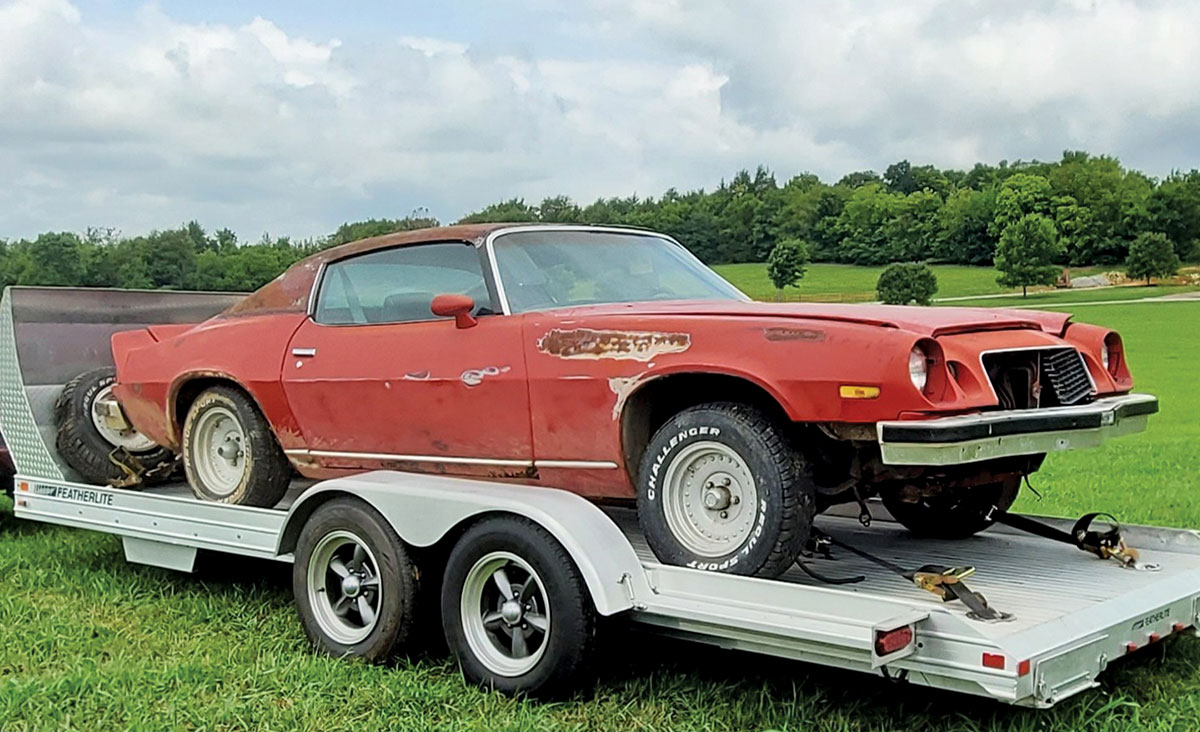 A rusted red classic car sits on a metal Featherlite trailer in a grassy field.
