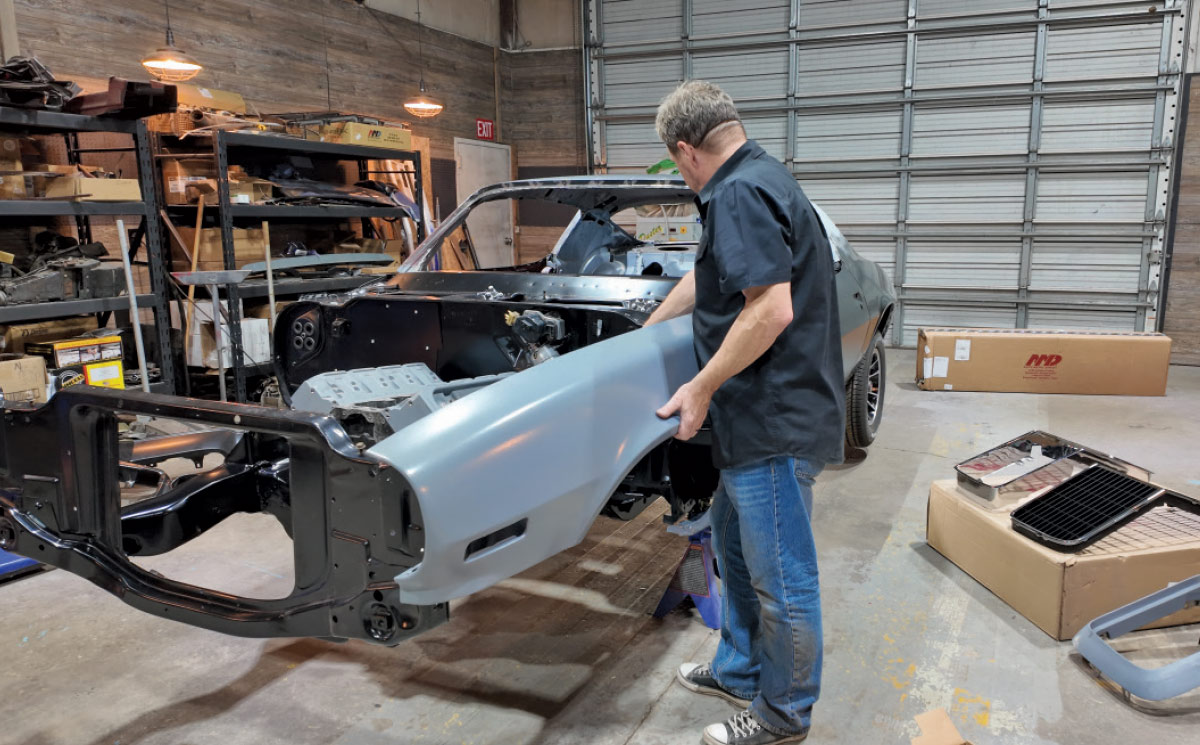 A mechanic aligns a blue primed fender onto the black front chassis of a classic car under restoration.