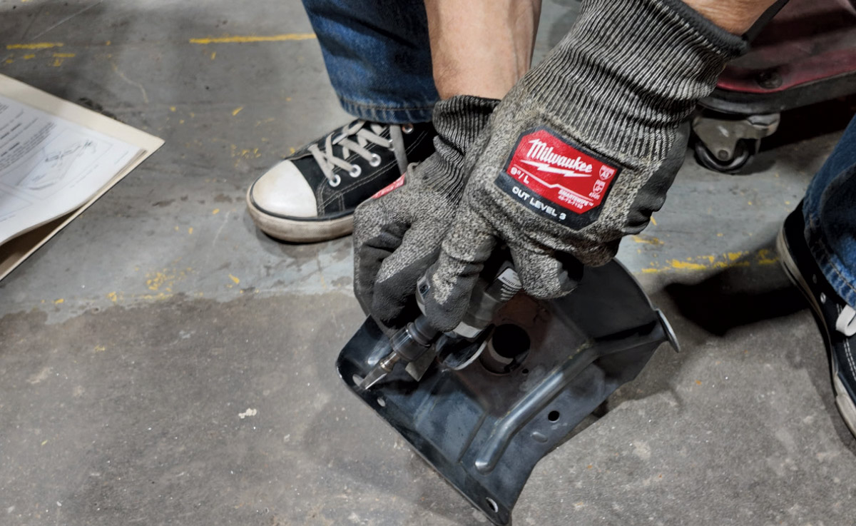 Hands in work gloves use a power tool to work on a black metal car part.