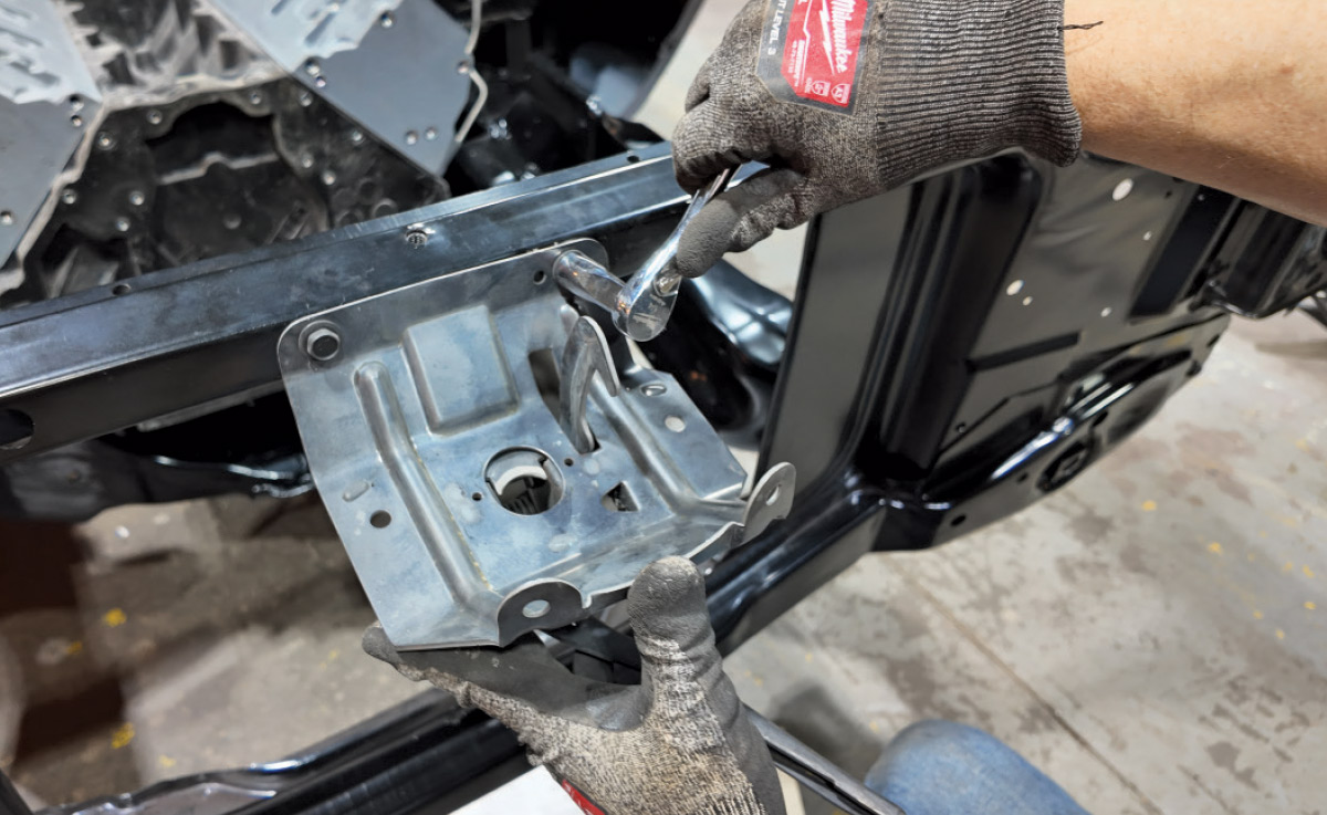 A person in work gloves uses a socket wrench to bolt a metal part onto a black car frame.