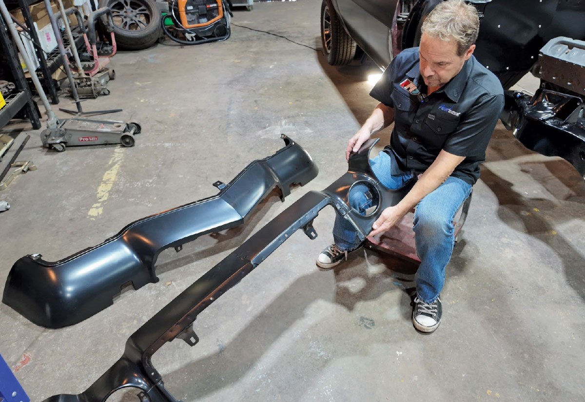 A mechanic sits on a workshop floor examining various black front-end car body parts.