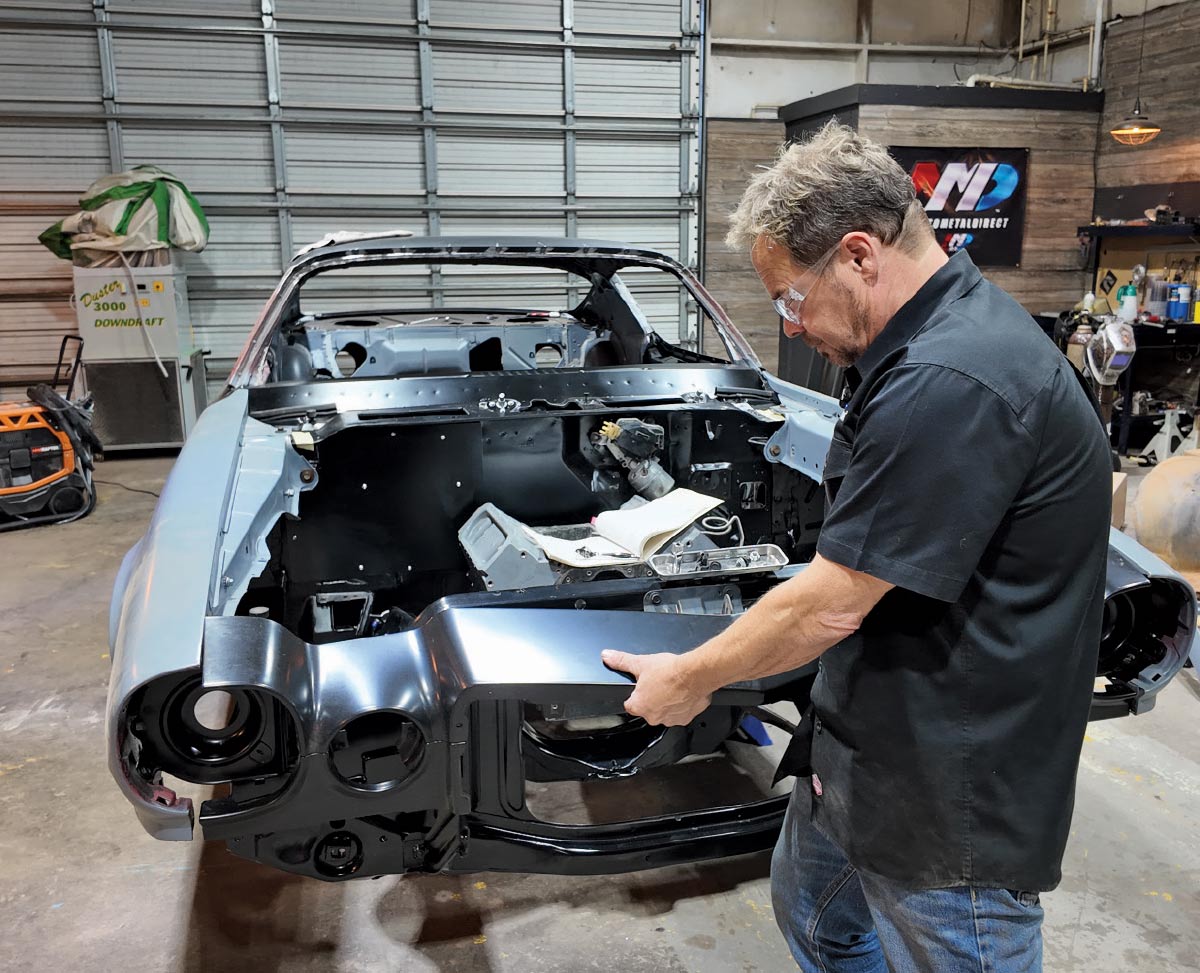 A mechanic wearing safety glasses test-fits a black front-end body piece onto a classic car.