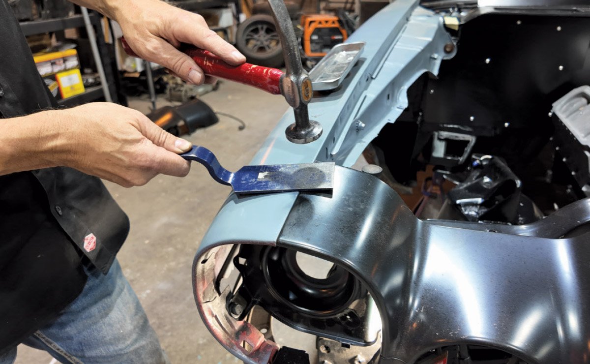 A mechanic uses a hammer and a blue pry tool to align metal panels on a car's front end.