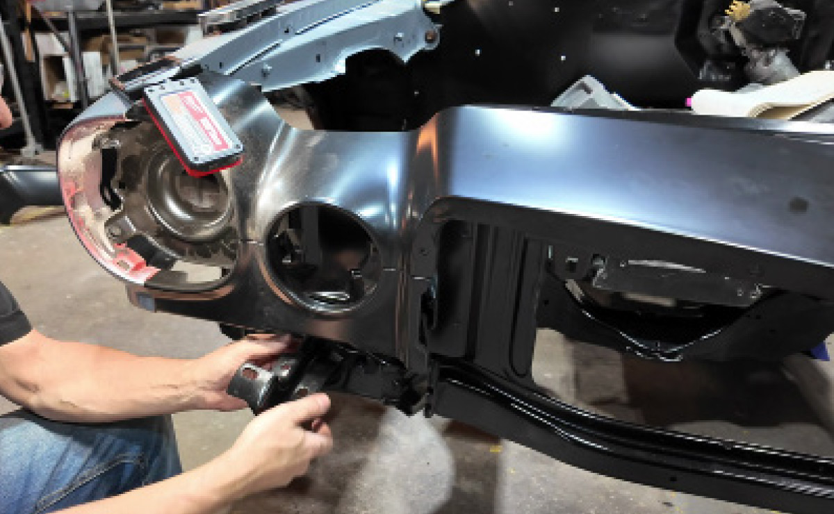 Hands maneuver a black metal structural component into place on a car chassis.