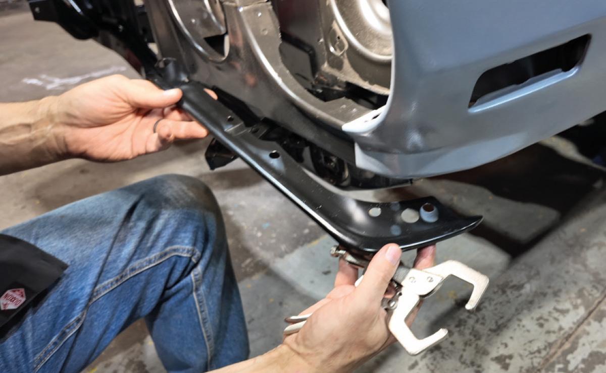 A person uses a metal clamp to hold a black trim piece in place on a car's front end.