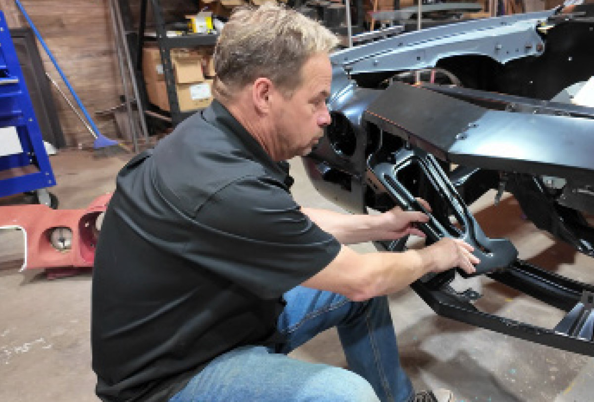 A mechanic kneels to install a black metal bracket onto a classic car's front chassis.