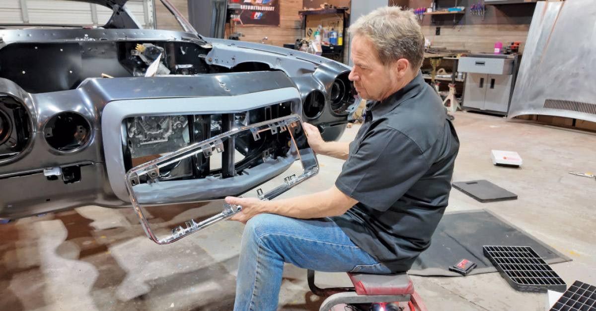 A mechanic sits and holds a chrome grill frame in front of a classic car's front end.